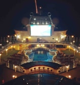 A luxury cruise ship deck featuring a pool surrounded by lounge chairs and warm ambient lighting. A large screen overlooks the area, showing an image against a dark night sky.