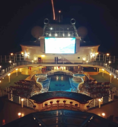A luxury cruise ship deck featuring a pool surrounded by lounge chairs and warm ambient lighting. A large screen overlooks the area, showing an image against a dark night sky.