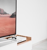 A modern interior setting featuring a white entertainment console with wooden accents. On the console, there is a gaming console with a controller on top. A large framed picture of a desert landscape with red sand and rocky formations is positioned next to the console.