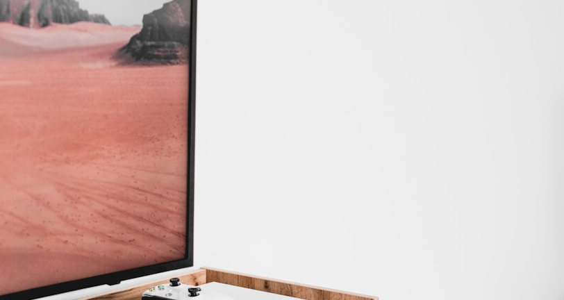 A modern interior setting featuring a white entertainment console with wooden accents. On the console, there is a gaming console with a controller on top. A large framed picture of a desert landscape with red sand and rocky formations is positioned next to the console.