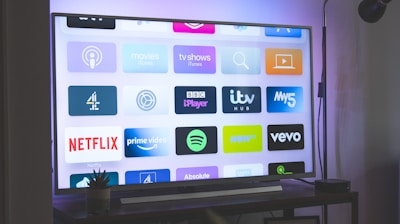 A flat-screen television is displaying a variety of streaming app icons, including Netflix, Prime Video, Spotify, and ITV Hub among others. The TV is placed on a media console with a small potted plant beside it. A tall lamp stands to the right, and a faint, ambient light casts a purple hue on the wall above the TV.