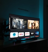 A television screen displaying a user interface with popular streaming service apps including Netflix, Sky, Apple TV, Prime Video, and others. The screen also shows images from movies or shows above the apps. The room is dimly lit with a potted plant visible on the side.