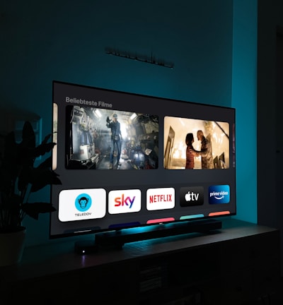 A television screen displaying a user interface with popular streaming service apps including Netflix, Sky, Apple TV, Prime Video, and others. The screen also shows images from movies or shows above the apps. The room is dimly lit with a potted plant visible on the side.