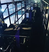 A narrow, industrial-looking platform with a series of large, black equipment cases secured with straps. Several control panels with glowing blue lights are visible on the front of each case. The platform is enclosed with metal railings, and various cables and chains hang along the sides, adding to the technical and utilitarian atmosphere. Bright lighting from behind casts a dramatic contrast against the darker sections of the platform.