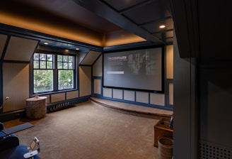 A cozy home theater room features a large projection screen displaying a movie selection interface. The room has dark wood paneling with soft, warm lighting highlighting the ceiling and walls. A large window is situated on one side with an outdoor view partially obscured by trees. There is a curved platform below the screen and a comfortable seating area, including a chevron-patterned ottoman. The room has a neutral color palette with beige, dark gray, and soft brown tones.