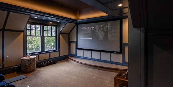 A cozy home theater room features a large projection screen displaying a movie selection interface. The room has dark wood paneling with soft, warm lighting highlighting the ceiling and walls. A large window is situated on one side with an outdoor view partially obscured by trees. There is a curved platform below the screen and a comfortable seating area, including a chevron-patterned ottoman. The room has a neutral color palette with beige, dark gray, and soft brown tones.