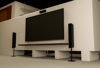 A modern home entertainment setup featuring a large flat-screen TV mounted on a light-colored wall. Two tall black speakers stand on either side of the TV. The room has a minimalist design with a clean, uncluttered look. Shelving and a wooden door are visible in the background.