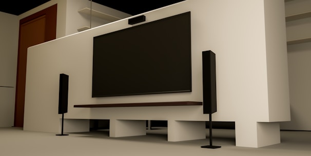 A modern home entertainment setup featuring a large flat-screen TV mounted on a light-colored wall. Two tall black speakers stand on either side of the TV. The room has a minimalist design with a clean, uncluttered look. Shelving and a wooden door are visible in the background.