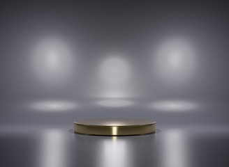 A metallic platform is centered in a minimalistic setting with a reflective surface. Soft, diffused lighting creates circular patterns on the background wall, enhancing the focus on the central stage. The ambiance is sleek and modern, with a hint of elegance.