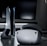 A sleek virtual reality headset sits on a reflective surface beside a gaming console and its controller. The background features a large dark monitor screen, creating a modern and futuristic setting. The lighting is subdued, highlighting the glossy surfaces of the electronic devices.