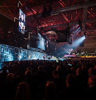 A large indoor event with a stage featuring bright screens and spotlights, attended by a substantial crowd. The venue has a high ceiling with industrial beams and red lighting accents. The atmosphere resembles a concert or a conference with a focus on visual effects.